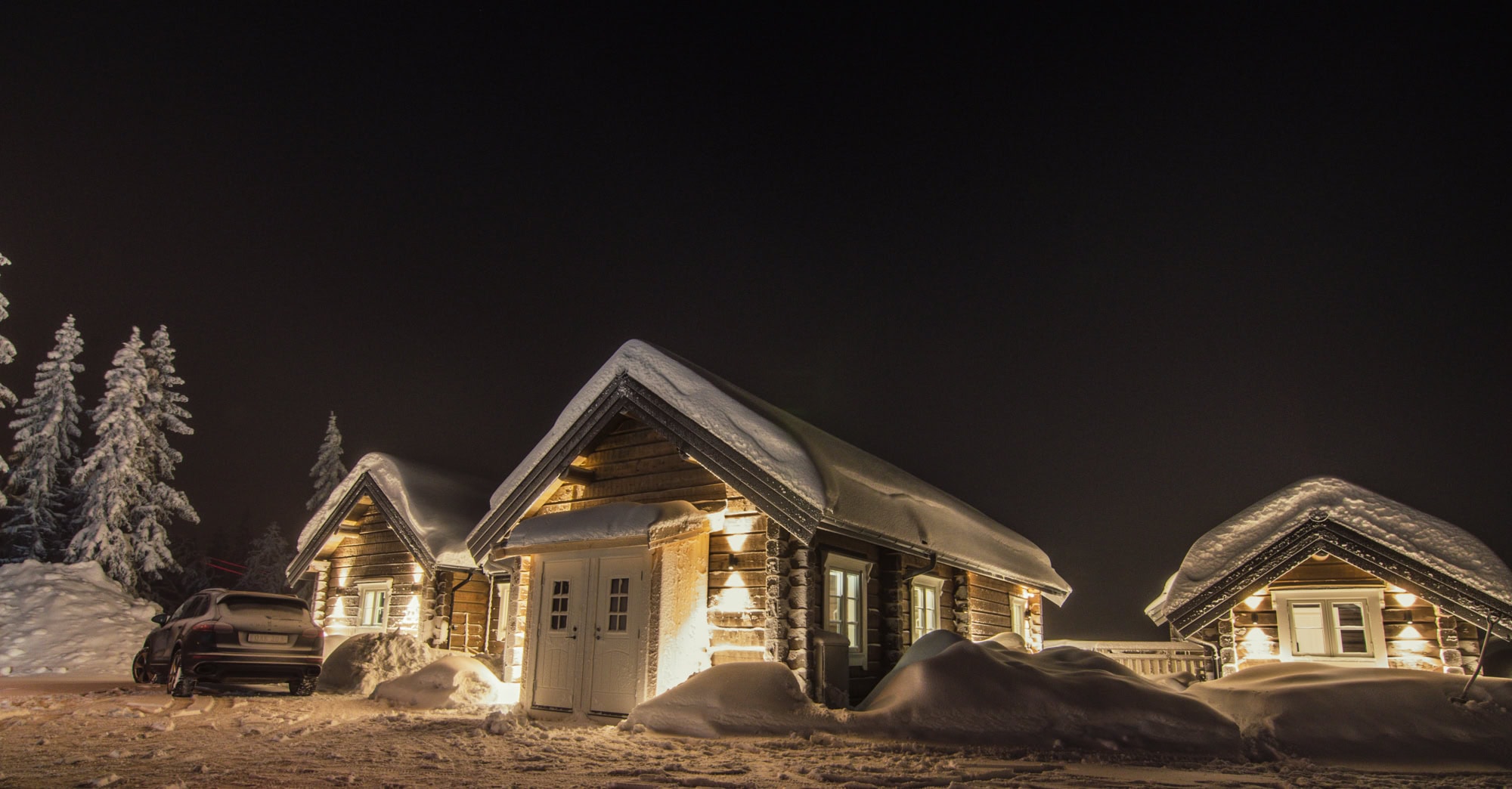 Ljusbelysta vedstuga i vinternatt med snötyngda tak och träd, skapar en mysig vinterkänsla.