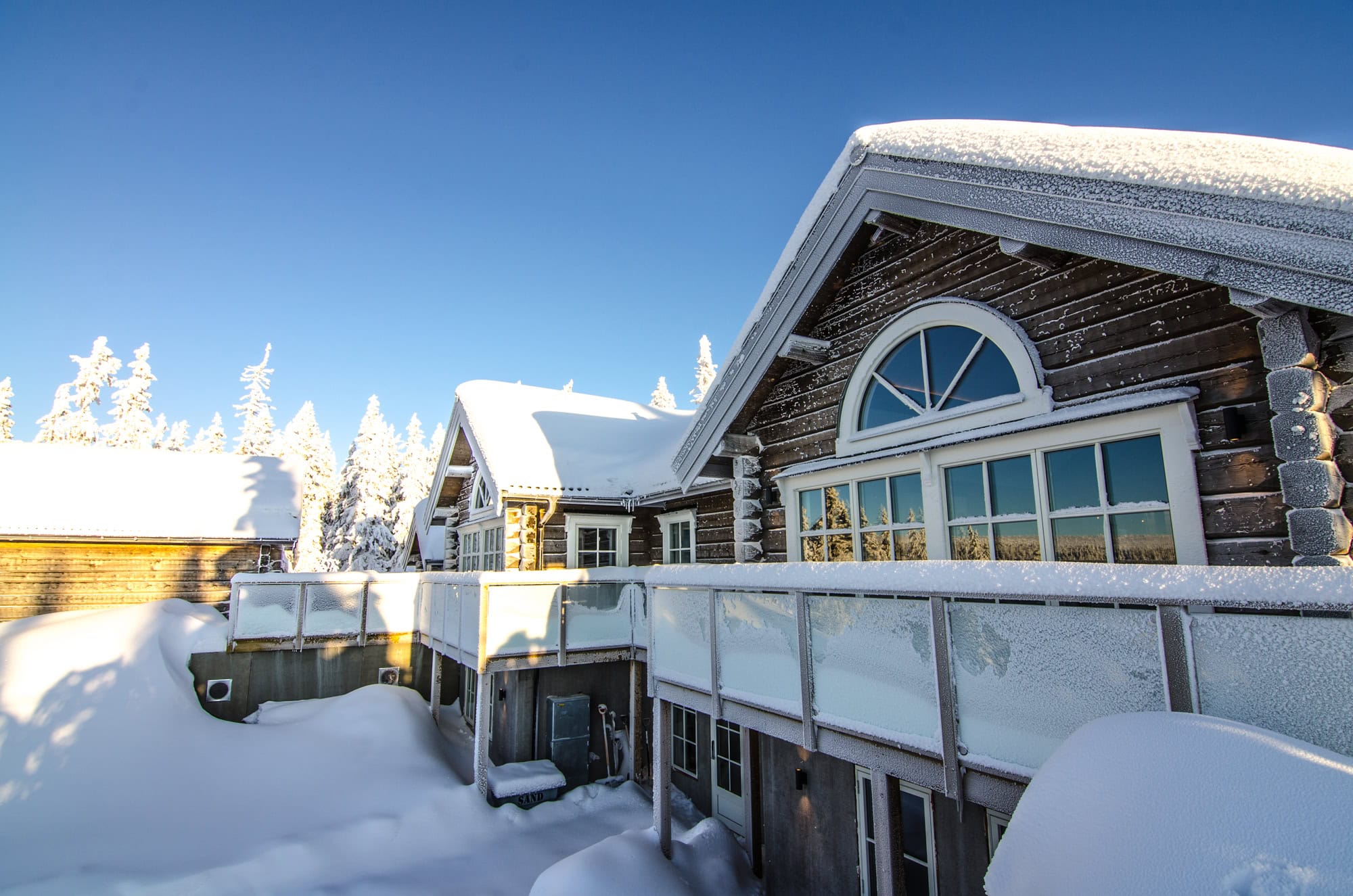 Snöklädd sommarstuga med stora fönster och balkong i vinterskrud, omgiven av snöiga träd och klar vinterhimmel. Perfekt för ett friluftsliv i kallt klimat med kvalitet även under vintersäsongen.