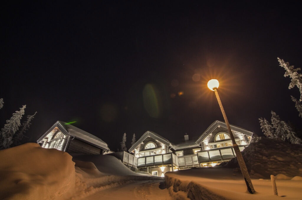 Snyggt belyst vinterhus under höstnatt med snö, kall vinter, ljus utomhusbelysning och snötäckta träd, perfekt för en mysig och varm vinterkåk i skandinavisk natur.