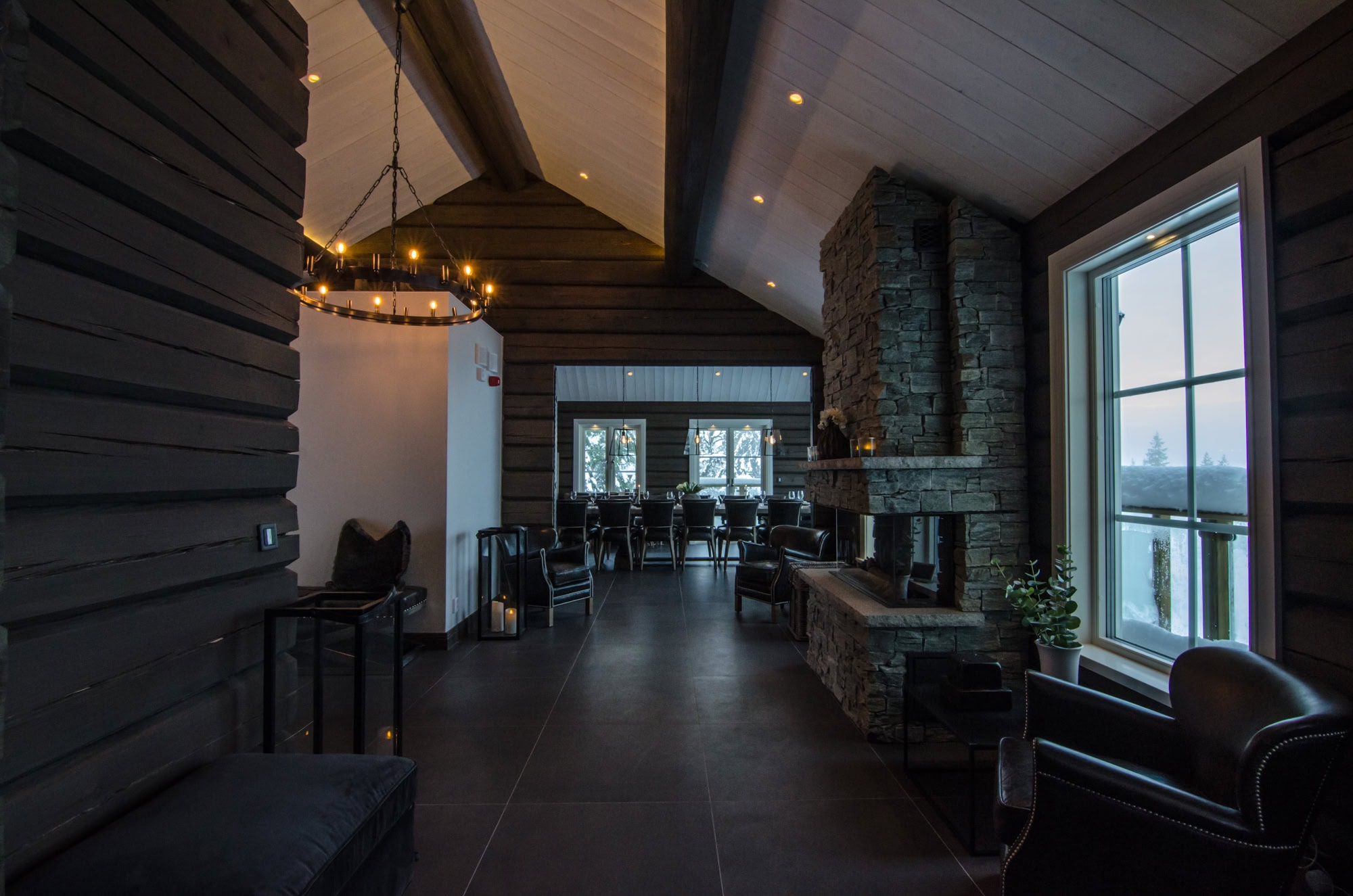 Vacker inomhusmiljö i en modern, rustik stuga med exklusiv inredning och stor öppen planlösning med braskamin, svarta stolar, mörka golv, stora fönster och mysig belysning.