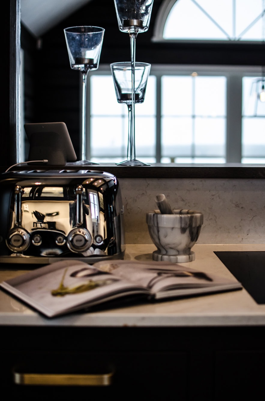 Skärbräda med recept och brödkniv, svart brödrost och marble mortar and pestle, tomt glas, avkopplande köksmiljö, naturligt ljus, moderna köksredskap för optimal matlagning och bakning.