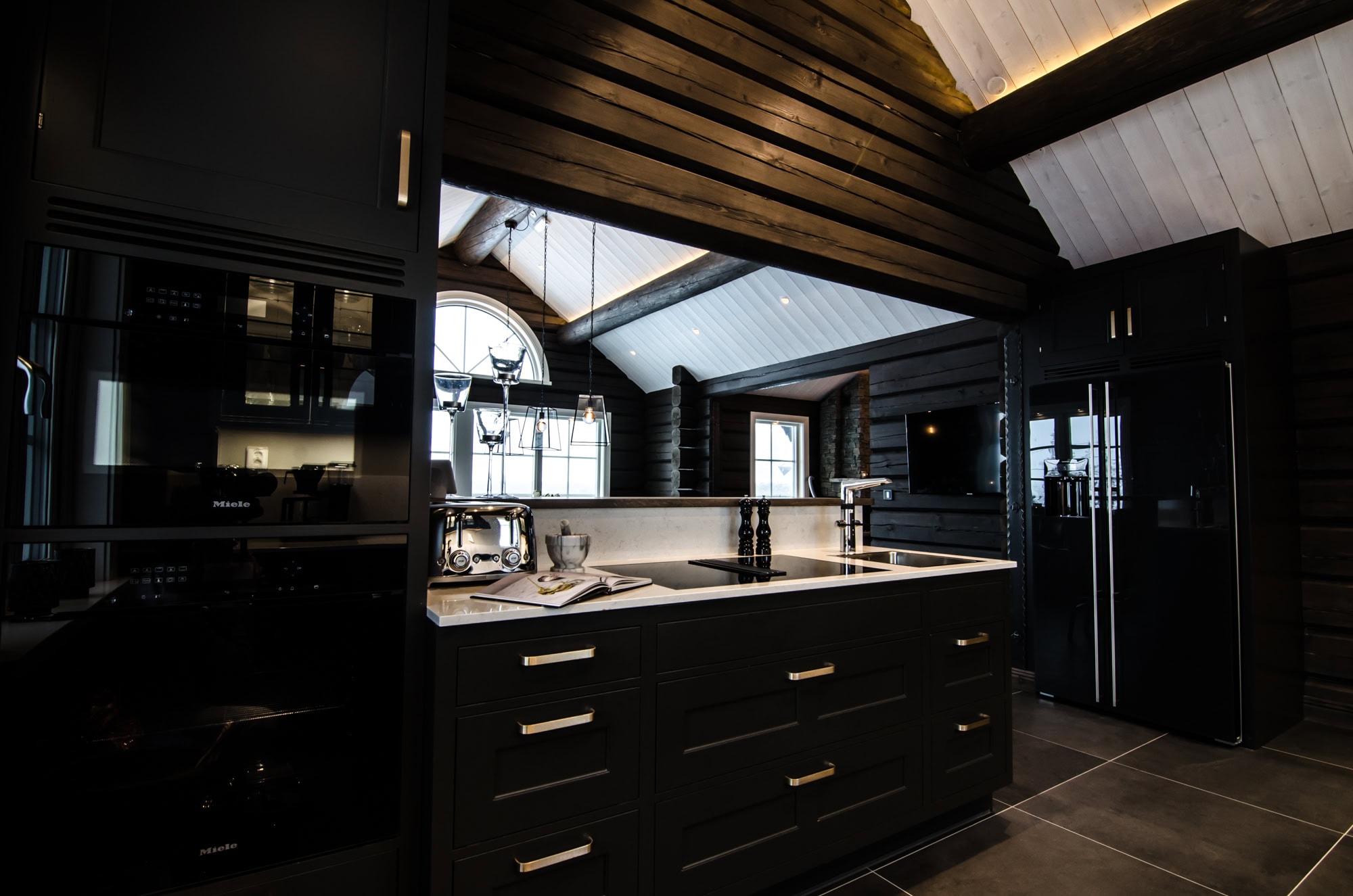 Elegant köksinredning med svart skåp och moderna vitvaror i rustik modern stuga. Naturligt ljus från fönster och takfönster skapar en inbjudande atmosfär för matlagning och samvaro.