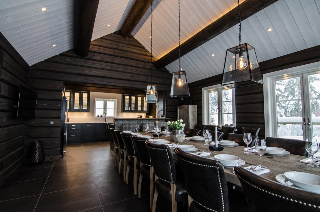 Elegant rustik matsal med svart träväggar och tak, stor träbord med vita tallrikar, glas, ljus, modern belysning, stora fönster med utsikt, perfekt för fest och familjemiddag.