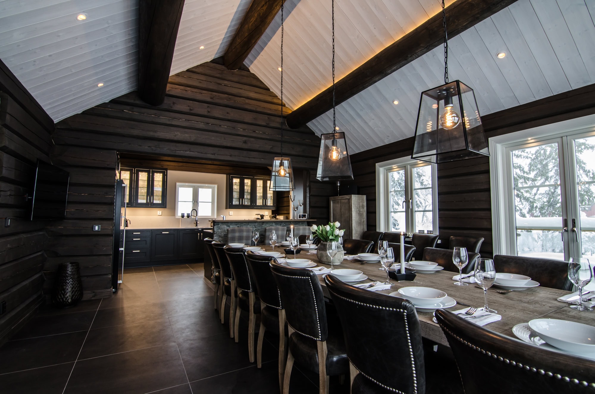 Elegant rustik matsal med svart träväggar och tak, stor träbord med vita tallrikar, glas, ljus, modern belysning, stora fönster med utsikt, perfekt för fest och familjemiddag.