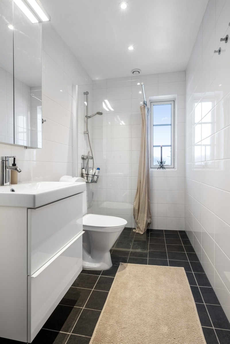 Modernt vitt badrum med dusch, toalett och handfat, mörk klinker och ljus matta, naturligt ljus från fönster, minimalistisk design, perfekt för relax och avkoppling.