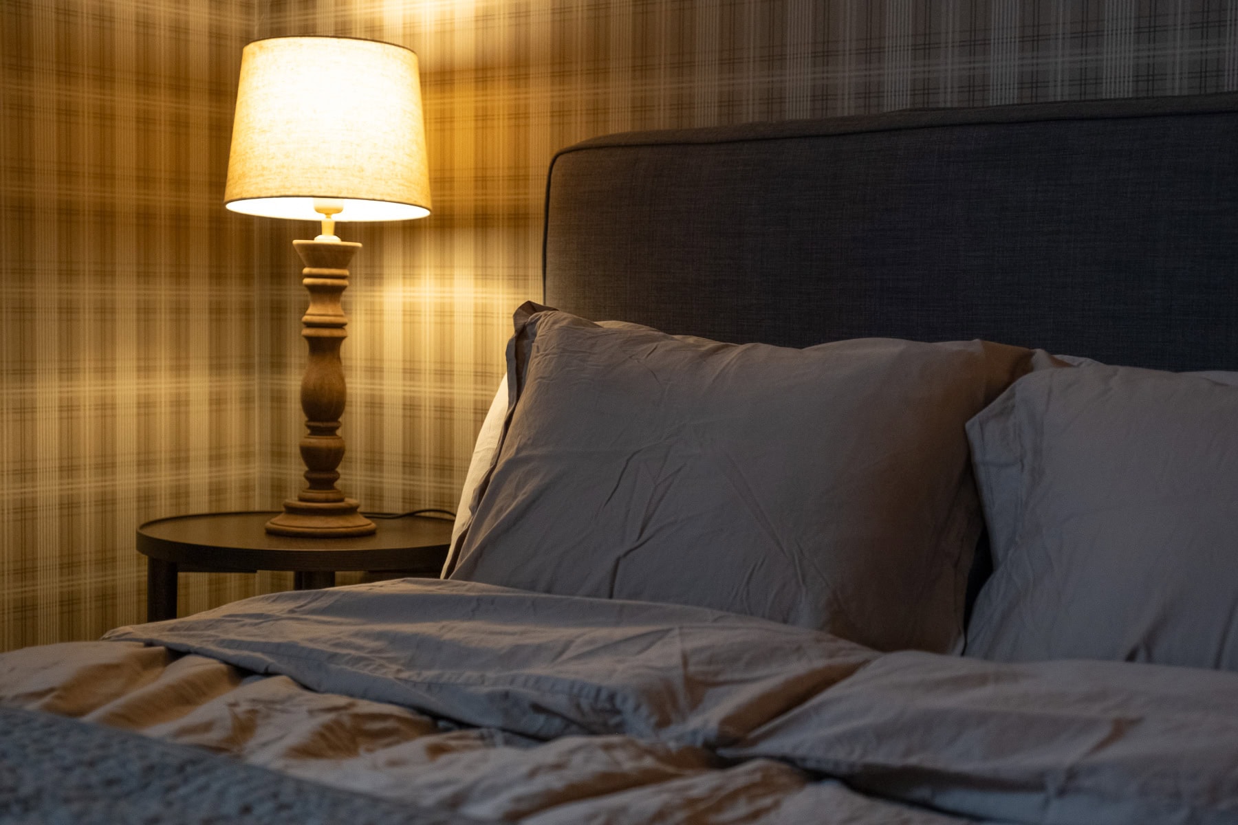 Mjuk säng med gråa kuddar i ett mysigt sovrum med varm belysning från bordslampa, perfekt för avkoppling och sömn.