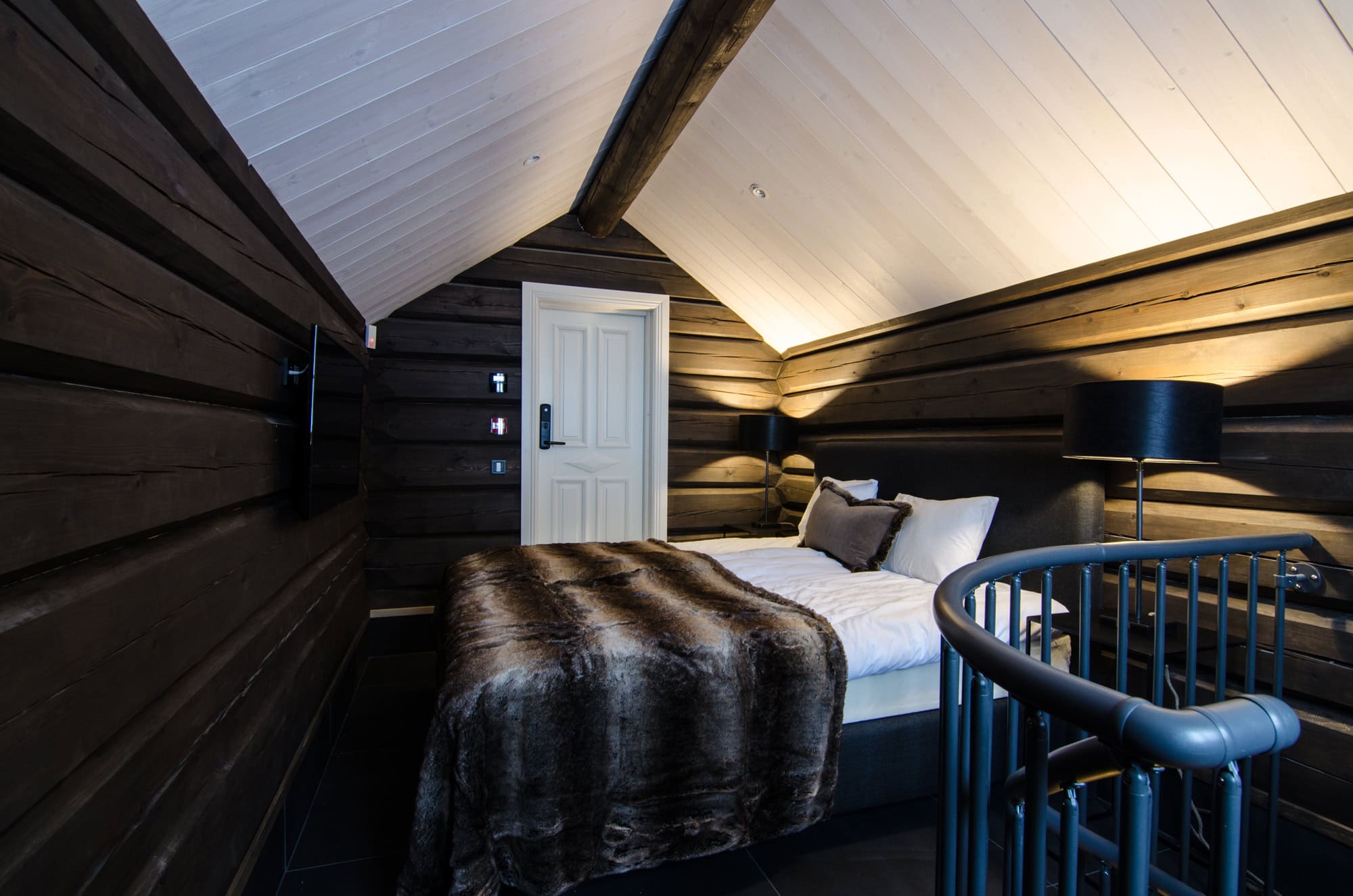 Elegant sovrum i rustik stil med mörka träväggar och tak, lyxig säng med pälsfilt, moderna lampor och ljudsystem, perfekt för ostörd vila och avkoppling i ett unikt trähus.