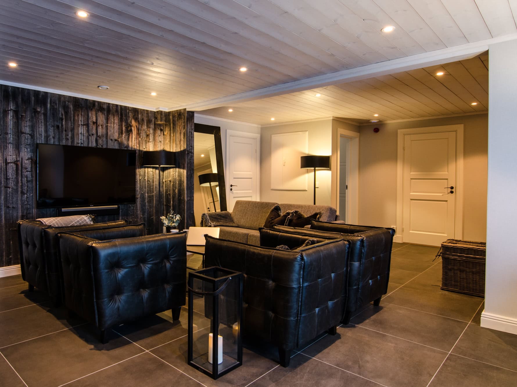 Modern inredning i gäststuga med komfortabla sittmöbler, svart soffa och TV, perfekt för avkoppling och umgänge. Trägolv och behaglig belysning skapar en inbjudande atmosfär.