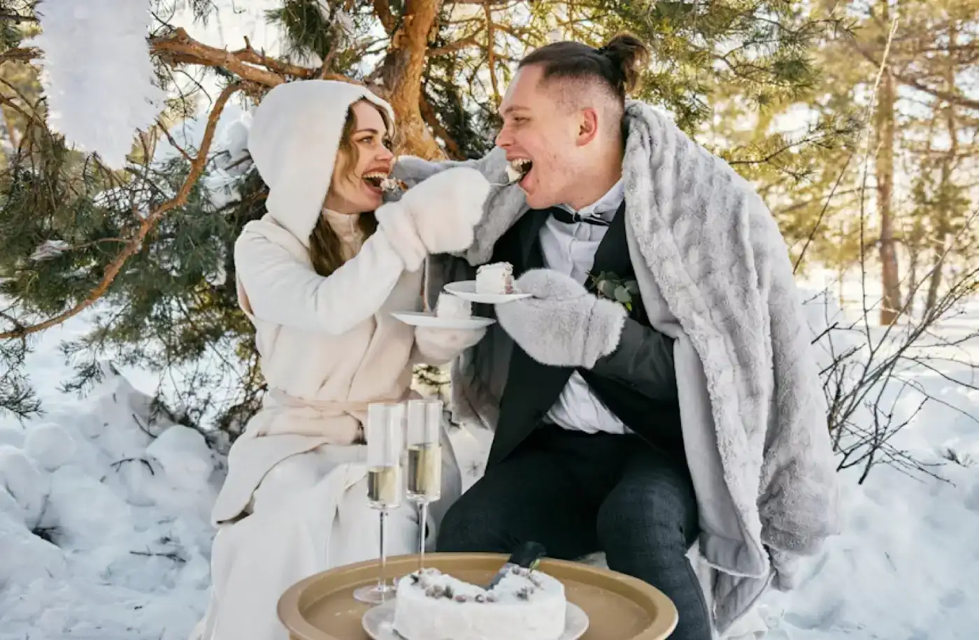 Brudeparet deler kake utendørs i snødekte fjellomgivelser, pakket inn i et teppe med champagneglass og vinterstemning.