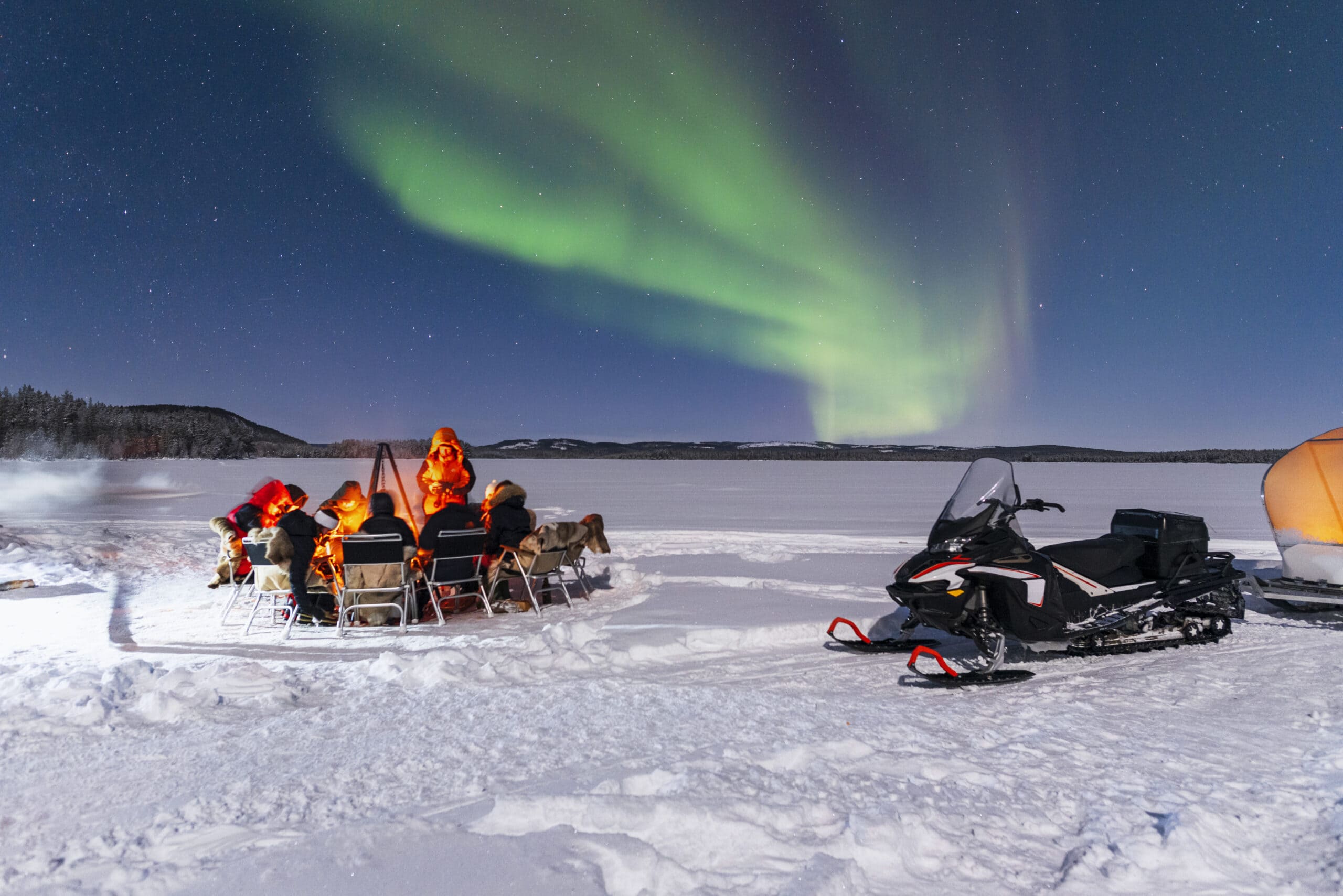 En grupp människor sitter runt en öppen eld på en frusen sjö i Sälen medan norrskenet lyser på himlen. En snöskoter står parkerad intill.