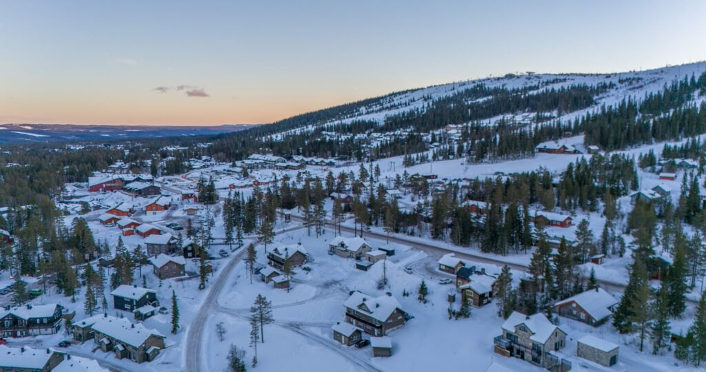 Snöklädd fritidsby i fjällmiljö med traditionella trähus, omgiven av skog och berg, perfekt för skidåkning och avkoppling i naturen.