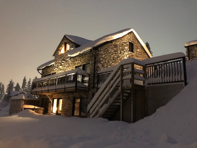 Großes Steinhaus in einer winterlichen Schneelandschaft, mit beleuchteten Fenstern und einer Außentreppe, umgeben von schneebedeckten Bäumen und der Landschaft.
