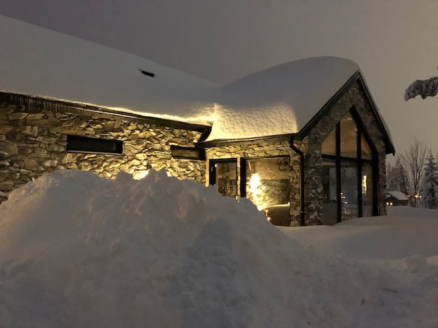 Schneebedeckte Hütten in einer Winterlandschaft mit großen Schneemassen und warmer Außenbeleuchtung.