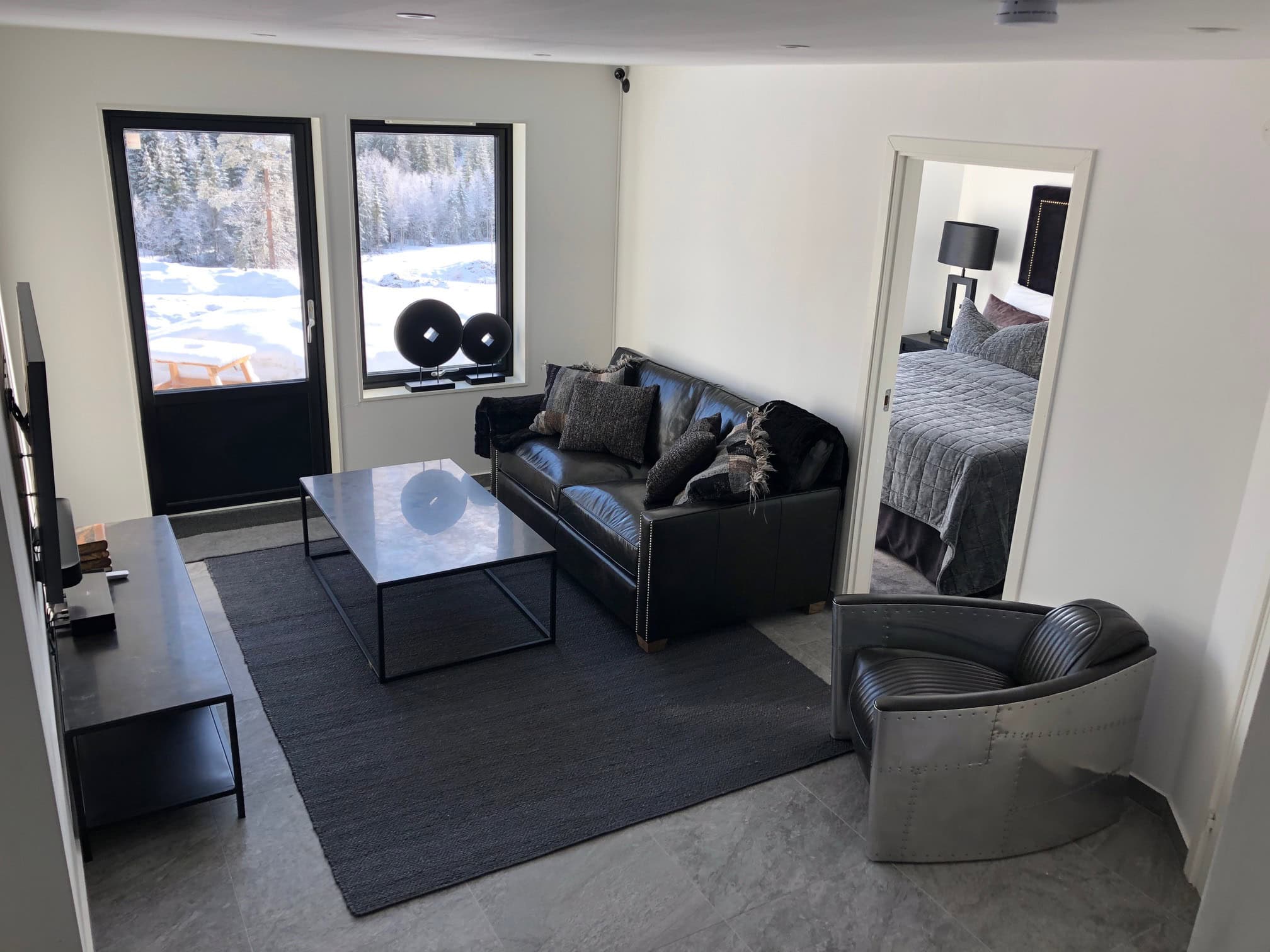 Modernes Wohnzimmer mit schwarzem Sofa, Sessel und großen Fenstern, durch die man die verschneite Landschaft draußen sehen kann.