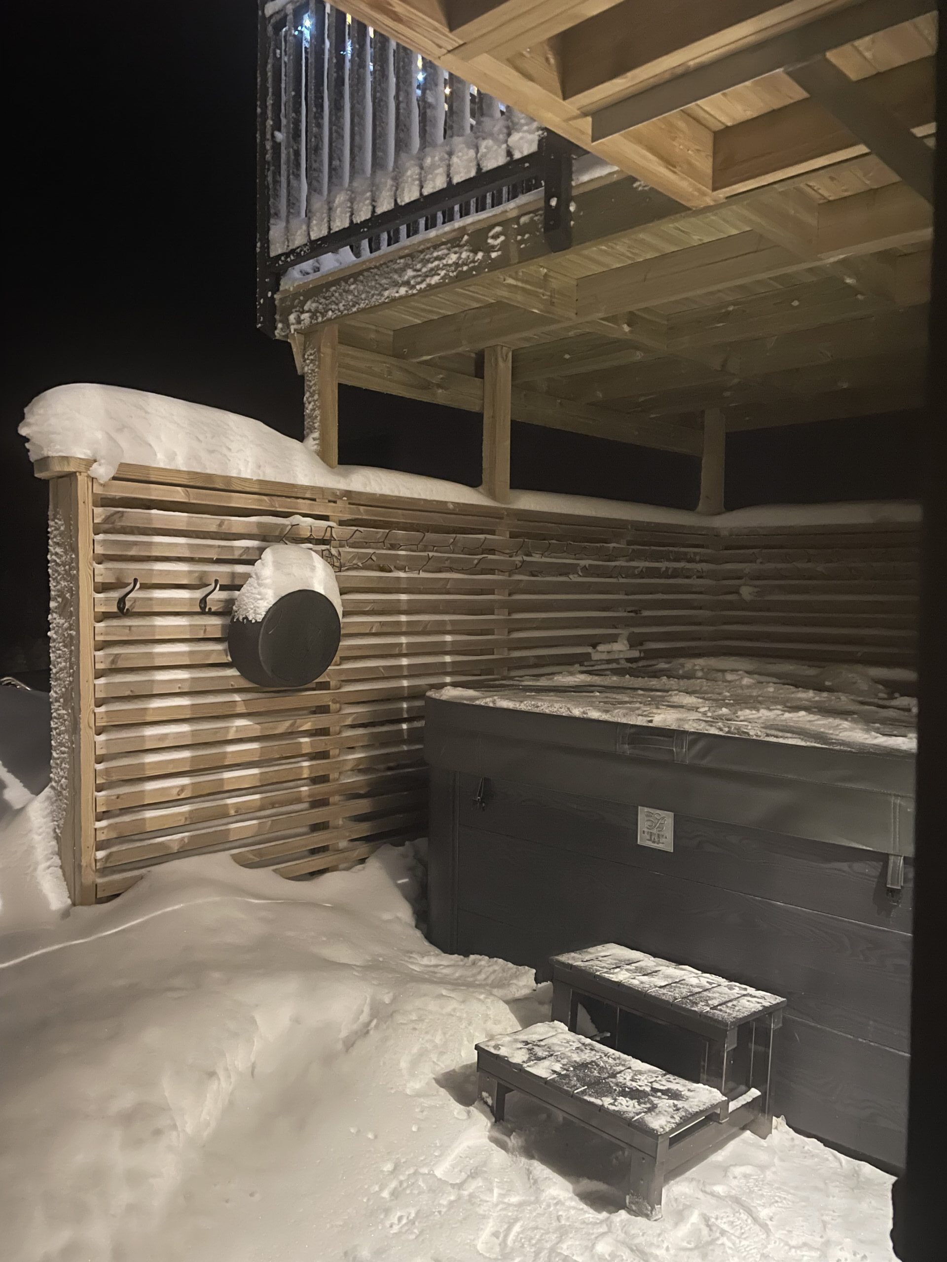 Blick auf eine verschneite Terrasse mit Holzzaun und Gartenmöbeln, darunter ein schwarzer Grill und eine Heizlampe, am Abend.