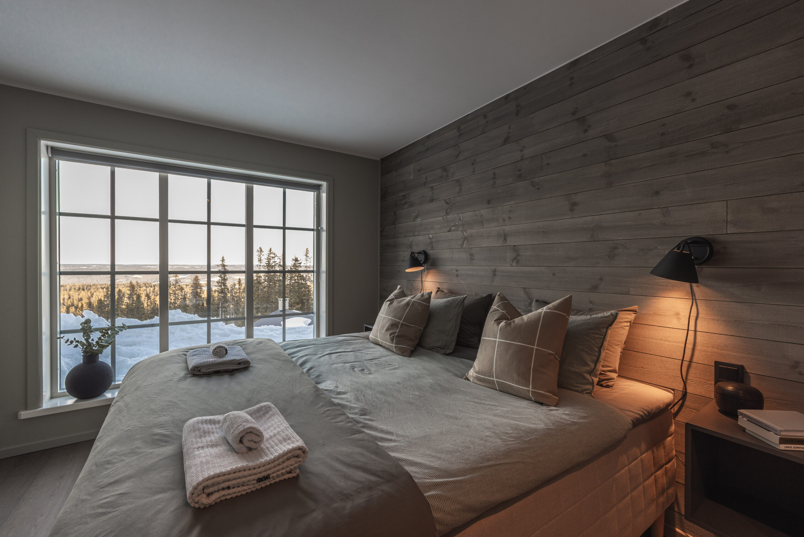 Mysigt sovrum med stor fönster som visar snöklädd landskap och skog, inrett med beige och gråa textilier, perfekt för avkoppling.