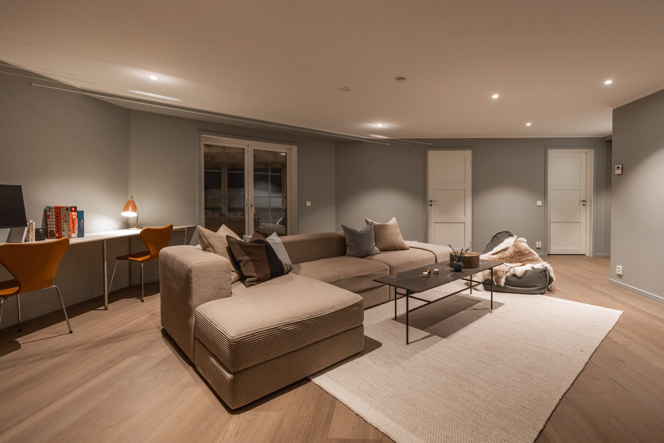 Wohnzimmer mit großem beigem Sofa, Arbeitsbereich mit Schreibtisch und Stühlen, hellgrauen Wänden und Holzböden, gemütlicher und moderner Einrichtung.