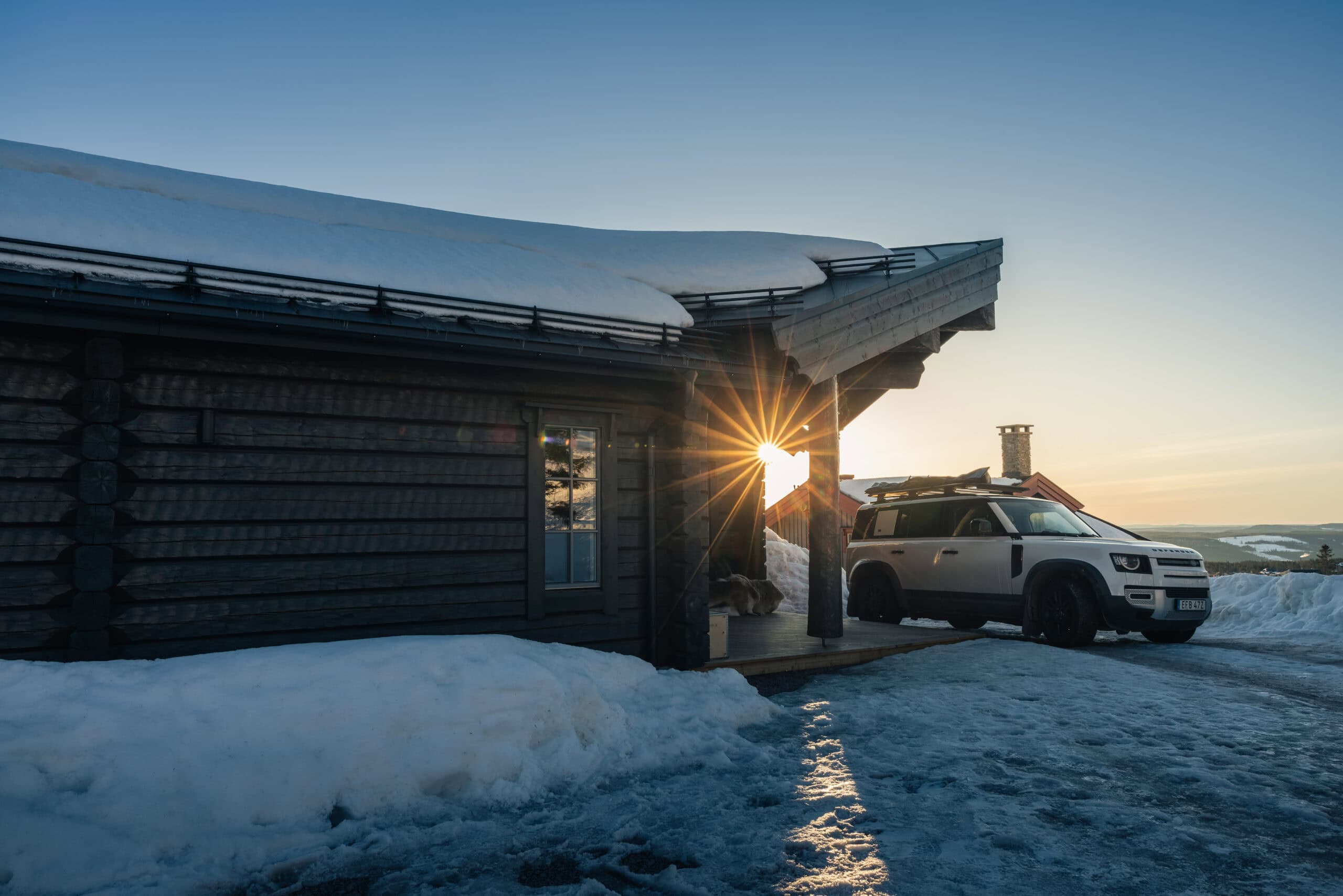 Salengodset stuga i snölandskap med bil parkerad utanför, soluppgång i bakgrunden, vintermiljö, fjäll och snö.