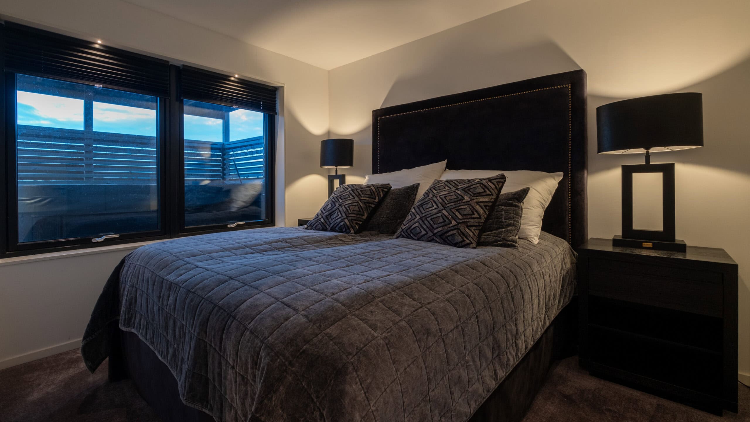 A bedroom with a double bed, bedside lamps on both sides and three windows.