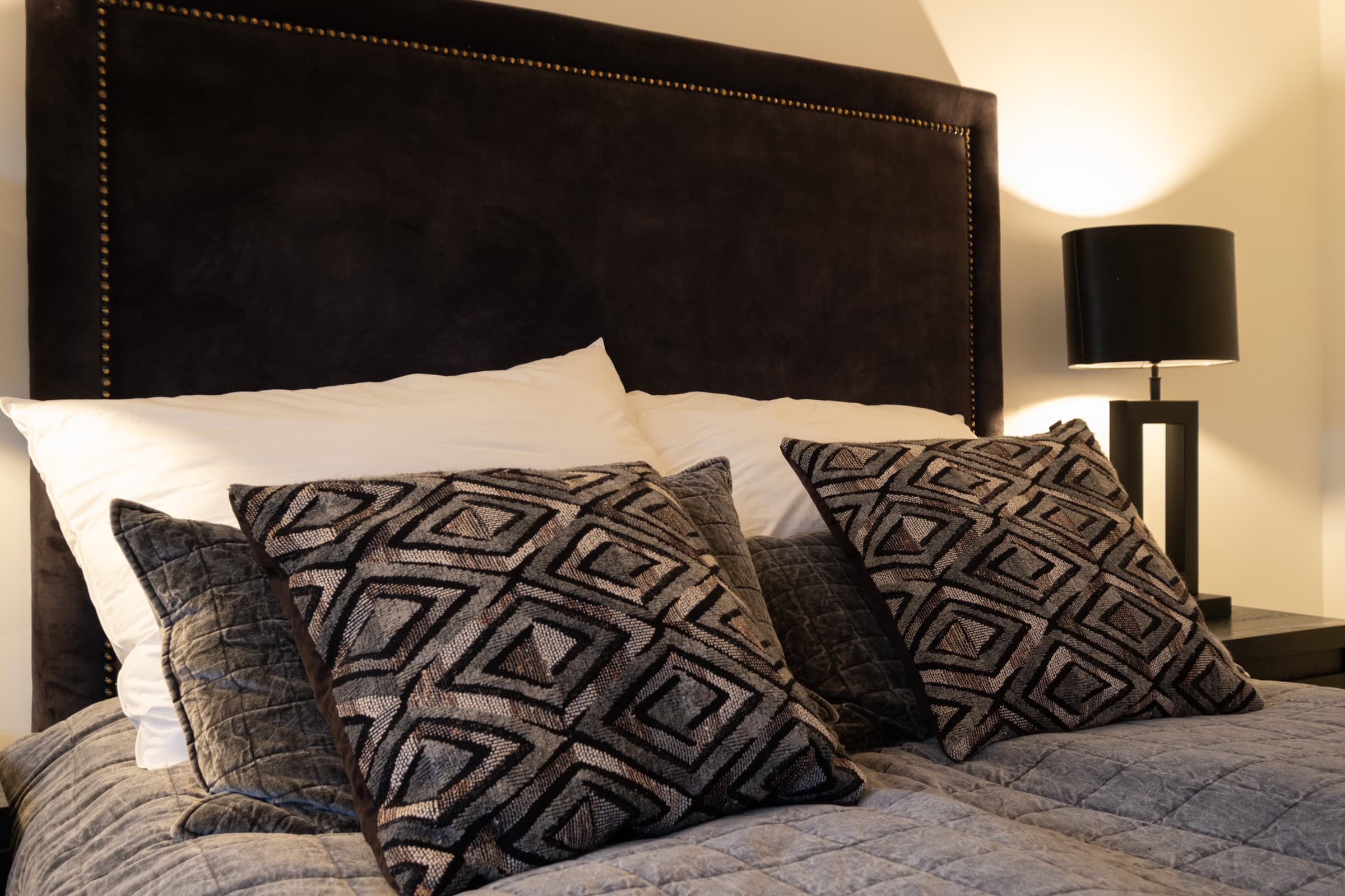 A headboard in dark leather with patterned cushions and a lit bedside lamp.
