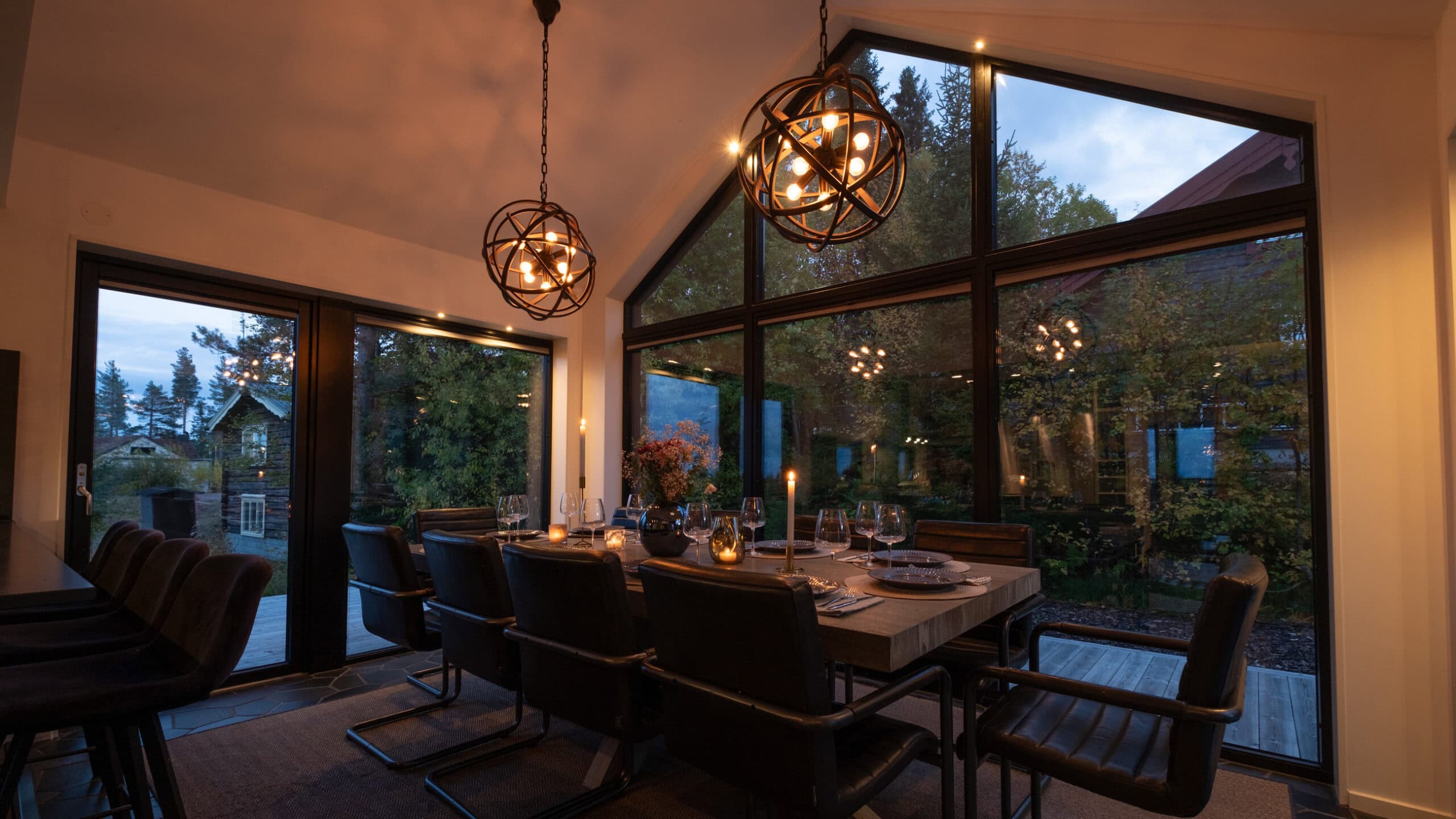 The long table set at dusk beneath the globe lamps, with the forest outside the windows.