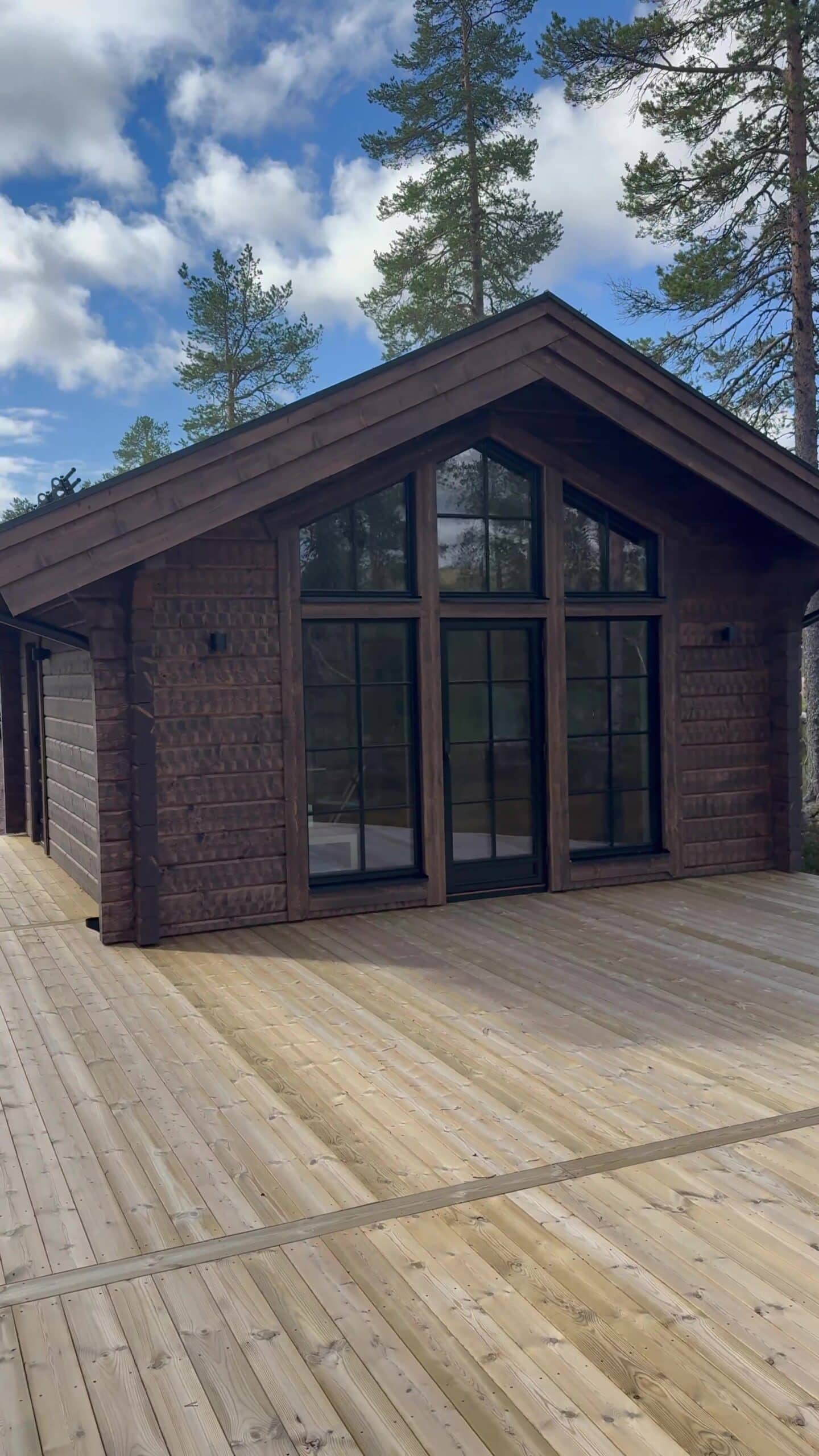 Die Giebelseite der Villa Åsstigen mit dunkler Holzvertäfelung, hohen Fenstern und Glastüren, die auf eine neu errichtete Holzterrasse führen, umgeben von einem Kiefernwald.